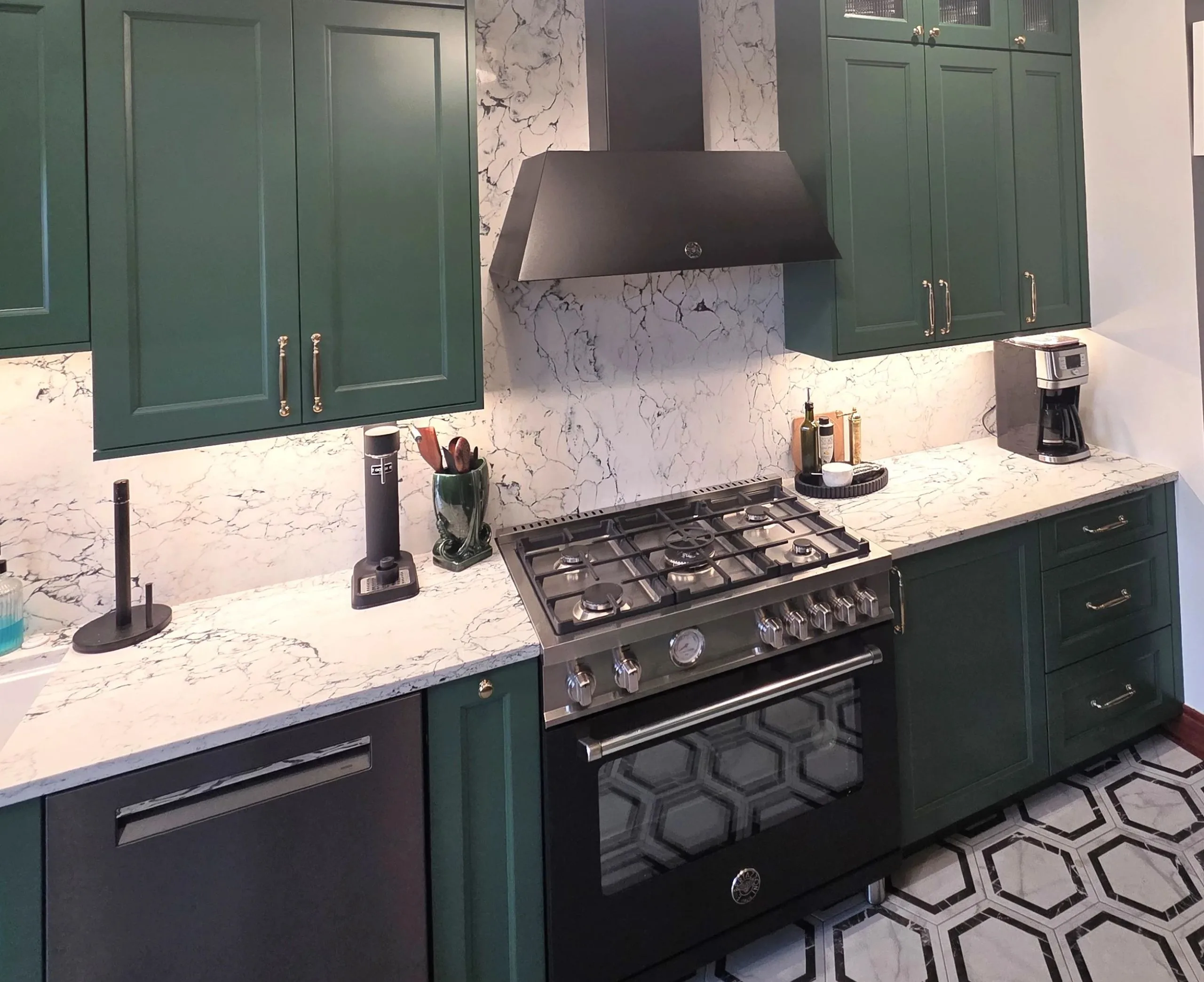 A luxury kitchen design by Maya Construction showcasing deep forest green shaker cabinets, a dramatic white marble-veined backsplash and countertop, and a high-end stainless steel gas range.